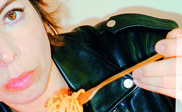 Photo de la chanteuse Mademoiselle K L’image montre une personne partiellement cadrée, principalement sur le côté gauche de l’image. Elle porte une veste en cuir noir avec des boutons métalliques. Elle tient une fourchette contenant des spaghettis. Le fond est clair et uni. La personne regarde vers l’objectif avec une expression neutre.