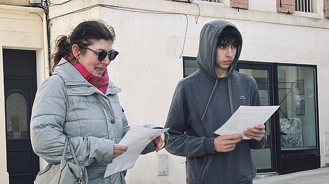Requête réussie Gemini said Cette photo montre deux personnes debout côte à côte sur ce qui semble être une place publique ou une zone piétonne pavée, devant un bâtiment aux murs clairs. Les Personnes À gauche : Une femme portant une doudoune grise, un foulard rose vif, des lunettes de soleil sombres et un sac à main gris en bandoulière. Elle tient des feuilles de papier et semble être en train de parler ou de lire à haute voix. À droite : Un jeune homme vêtu d'un sweat à capuche gris foncé (la capuche est relevée) et d'un pantalon de jogging assorti. Il tient également des feuilles de papier et regarde droit devant lui avec un air sérieux. Le Décor Arrière-plan : Le bâtiment derrière eux est de style européen classique avec des volets en bois (peints en rose délavé) et une vitrine de magasin au rez-de-chaussée. Sol : Le sol est constitué de larges dalles de pierre claire, typiques des centres-villes historiques ou des esplanades.