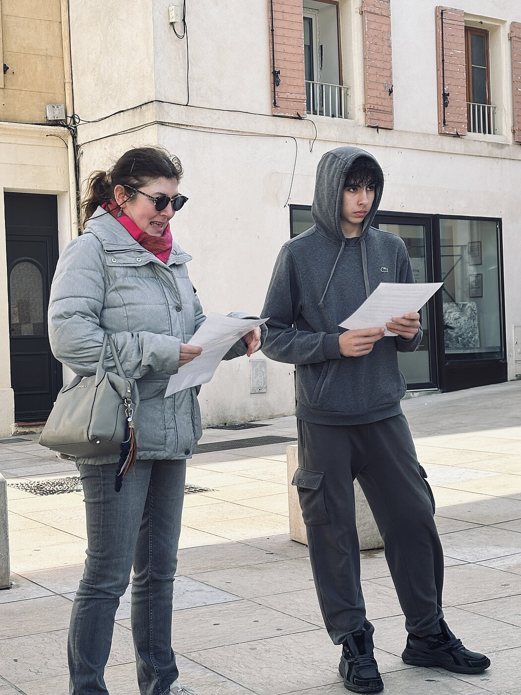     Requête réussie  Gemini said  Cette photo montre deux personnes debout côte à côte sur ce qui semble être une place publique ou une zone piétonne pavée, devant un bâtiment aux murs clairs. Les Personnes      À gauche : Une femme portant une doudoune grise, un foulard rose vif, des lunettes de soleil sombres et un sac à main gris en bandoulière. Elle tient des feuilles de papier et semble être en train de parler ou de lire à haute voix.      À droite : Un jeune homme vêtu d'un sweat à capuche gris foncé (la capuche est relevée) et d'un pantalon de jogging assorti. Il tient également des feuilles de papier et regarde droit devant lui avec un air sérieux.  Le Décor      Arrière-plan : Le bâtiment derrière eux est de style européen classique avec des volets en bois (peints en rose délavé) et une vitrine de magasin au rez-de-chaussée.      Sol : Le sol est constitué de larges dalles de pierre claire, typiques des centres-villes historiques ou des esplanades.