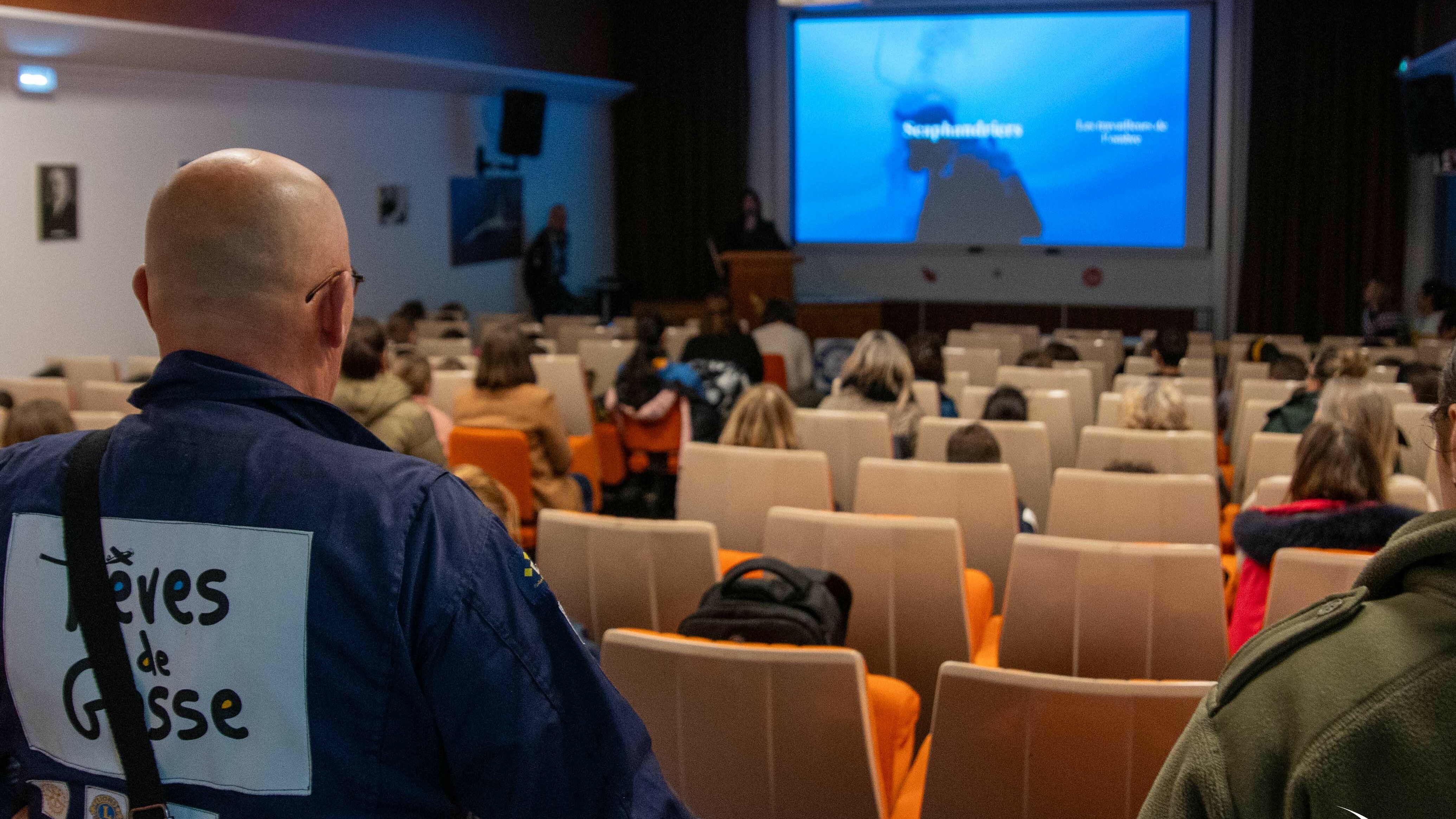     Requête réussie  Cette image montre une conférence ou une présentation se déroulant dans un auditorium ou une salle de projection.  Voici une description détaillée des éléments visibles : Premier plan      Un homme vu de dos, portant une combinaison bleue, occupe le côté gauche de l'image.      Sur son dos, un grand écusson blanc porte l'inscription "Rêves de Gosse" accompagnée d'un dessin d'avion stylisé.