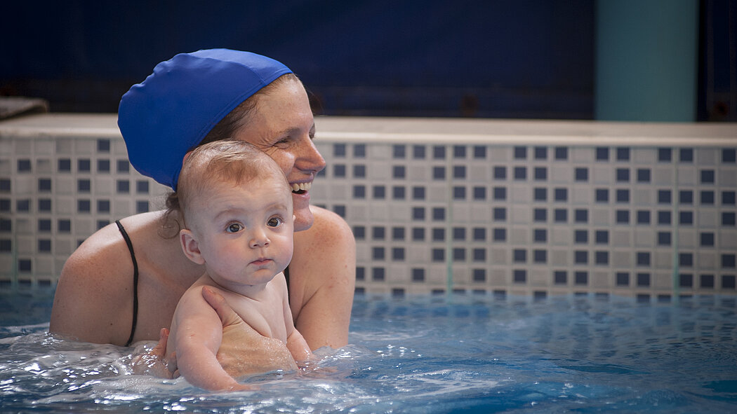 L’image montre deux personnes dans une piscine :      Une personne adulte porte un bonnet de bain bleu et est partiellement immergée dans l’eau. Elle tient un bébé dans ses bras.      Le bébé est nu (sans vêtements visibles) et également partiellement immergé. Il regarde droit devant lui.      En arrière-plan, on voit un mur recouvert de carreaux en mosaïque bleus et blancs.      L’eau de la piscine est bleue et on aperçoit quelques vaguelettes à la surface.