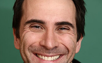 photo du comédie Thomas Poitevin L’image montre un portrait en gros plan d’un homme souriant. Il a les cheveux bruns, courts, et un front dégagé. Son visage est marqué de rides d'expression autour des yeux et de la bouche. Il a les dents visibles en souriant, les yeux plissés, et un léger début de barbe. L’arrière-plan est de couleur vert uni.