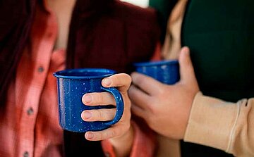 L'image montre deux mains tenant des tasses bleues en émail. La première tasse est saisie par une main portant un bracelet, tandis que la seconde est tenue par une autre main. Les tasses sont de couleur bleue avec des éclats blancs, typiques du style émaillé. En arrière-plan, on peut distinguer des vêtements : une chemise rose à carreaux et un gilet en doudoune verte, suggérant un environnement extérieur ou une ambiance chaleureuse. Les tasses sont tenues près du corps de chaque personne, mais les visages ne sont pas visibles dans cette image.