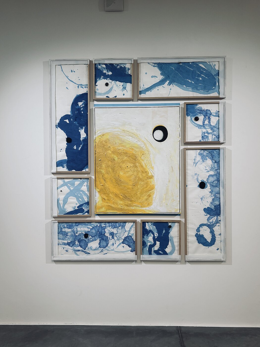 Cette œuvre est une composition murale multi-panneaux qui allie peinture abstraite et assemblage géométrique. Elle se compose d'un panneau central entouré de huit cadres rectangulaires de tailles variées, créant une structure globale équilibrée mais dynamique.  Voici une description détaillée de ses composants :  ### Le Panneau Central  * **Contenu :** Il présente une forme organique jaune ocre, texturée et dense, qui occupe la majeure partie de la surface blanche. On y voit également un cercle noir parfait, entouré d'un fin liseré blanc et noir, rappelant une éclipse ou un œil. * **Texture :** La peinture jaune semble appliquée en couches épaisses, créant un relief qui contraste avec la planéité des éléments environnants.  ### Les Cadres Périphériques  * **Style :** Huit cadres en bois clair entourent le centre. Ils contiennent des compositions à l'encre ou à l'aquarelle bleue sur fond blanc. * **Motifs :** Le bleu est appliqué de manière fluide, avec des effets d'éclaboussures, de taches et de lignes sinueuses évoquant des formes aquatiques ou cellulaires. * **Points Noirs :** Presque chaque panneau satellite contient un petit disque noir plein, créant un rappel visuel avec le cercle du panneau central et guidant l'œil à travers toute la structure.  ### Composition et Matériaux  * **Palette :** L'œuvre repose sur un contraste primaire entre le **jaune solaire**, le **bleu profond** et des touches de **noir**, le tout sur un fond blanc éclatant. * **Structure :** L'agencement des cadres rappelle une grille déconstruite. L'espace vide entre les cadres fait partie intégrante de l'œuvre, laissant respirer chaque élément.  L'ensemble dégage une impression de cosmologie ou de cartographie abstraite, où des éléments micro (les taches d'encre) et macro (la forme jaune centrale) coexistent. 