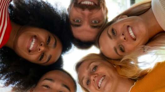 Sur cette image, cinq personnes sont visibles, leurs têtes rapprochées en cercle et regardant vers le bas, en direction de l’objectif de la caméra placé en dessous d’eux. Elles sourient toutes. La photo est prise en contre-plongée avec un effet de perspective circulaire. Chacune des personnes porte un vêtement de couleur différente (rouge rayé, vert, blanc, jaune, etc.). En arrière-plan, on distingue une grande fenêtre laissant passer la lumière naturelle.