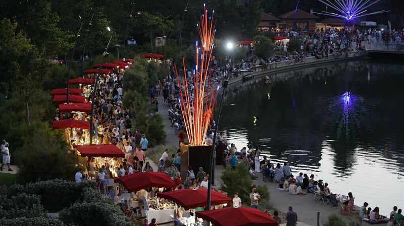 Un grand nombre de personnes rassemblées le long d’un plan d’eau.  Un chemin piéton bordé d’étals ou stands couverts de parasols rouges.  Des lumières artificielles illuminent les stands et une structure verticale composée de tiges rouges et bleues lumineuses.  Sur la rive opposée, une autre structure lumineuse en forme d’éventail ou d’étoile, émettant de la lumière violette.  Des arbres et végétation bordent les deux côtés du plan d’eau.  De nombreuses personnes sont assises ou debout au bord de l’eau.  L’éclairage suggère que la scène se déroule en fin de journée ou de nuit.