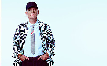 Photo de Gaetan Roussel On voit une personne photographiée en pied jusqu’au haut des cuisses, debout face à l’appareil. Elle porte une casquette noire.  Elle est vêtue d’une chemise blanche, d’une cravate claire avec de fines bandes colorées, et d’une veste à manches longues à rayures verticales noires et blanches.  Le pantalon est foncé. Les mains sont placées dans les poches du pantalon.  Le visage est visible, sans barbe. Le regard est dirigé vers l’avant.  Le fond de l’image est uni et très clair, proche du blanc ou du bleu très pâle, sans élément visible. L’éclairage est uniforme, sans ombre marquée.