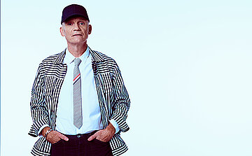 Photo de Gaetan Roussel On voit une personne photographiée en pied jusqu’au haut des cuisses, debout face à l’appareil. Elle porte une casquette noire.  Elle est vêtue d’une chemise blanche, d’une cravate claire avec de fines bandes colorées, et d’une veste à manches longues à rayures verticales noires et blanches.  Le pantalon est foncé. Les mains sont placées dans les poches du pantalon.  Le visage est visible, sans barbe. Le regard est dirigé vers l’avant.  Le fond de l’image est uni et très clair, proche du blanc ou du bleu très pâle, sans élément visible. L’éclairage est uniforme, sans ombre marquée.