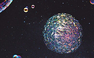 L’image montre une composition visuelle représentant un fond sombre parsemé de petits points lumineux rappelant des étoiles. Au centre, une grande sphère texturée et multicolore occupe une place proéminente. Elle présente des reflets irisés et une surface complexe. Plusieurs petites bulles ou sphères colorées flottent autour, créant un effet spatial ou cosmique. L’ensemble de l’image évoque un environnement abstrait ou inspiré de l’espace.