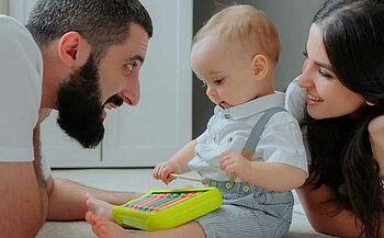 L'image montre trois personnes allongées ou assises sur un sol clair à l'intérieur d'une pièce. Un enfant est au centre, tenant un petit maillet et touchant un xylophone en plastique vert aux lames colorées. L'enfant porte une chemise blanche et une salopette rayée. Deux adultes sont placés de chaque côté de l'enfant, tous les deux souriants et tournés vers lui. En arrière-plan, on aperçoit des éléments de mobilier de couleur blanche.