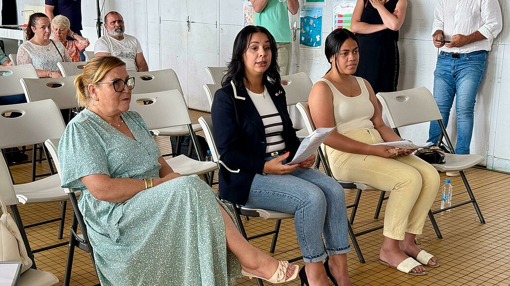 La photo montre un groupe de personnes assises sur des chaises pliantes dans une pièce carrelée aux murs blancs. Au premier plan, trois femmes sont assises sur la première rangée. 