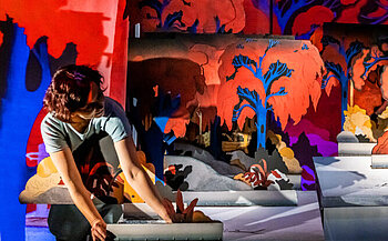 L’image montre une personne penchée vers l’avant, les mains posées sur une maquette ou un élément de décor. Elle porte un t-shirt bleu clair. À l’arrière-plan se trouvent plusieurs panneaux colorés représentant un paysage avec des arbres rouges, bleus et orangés sur un fond sombre, créant un contraste visuel fort. L’éclairage met en valeur les couleurs vives et les ombres, donnant une atmosphère théâtrale à la scène.