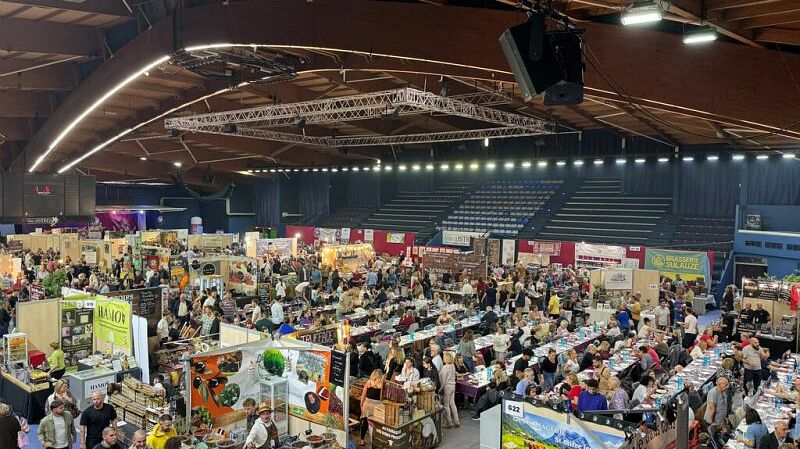 Cette image montre une étagère remplie de pots de confitures, de sauces et d'autres produits artisanaux. Les pots sont joliment disposés, avec des étiquettes colorées visibles sur chaque produit. On remarque une variété de couleurs, allant des rouges aux jaunes, représentant probablement des saveurs comme la fraise, l'abricot ou d'autres fruits. L'étagère est en bois, créant une ambiance rustique, et des éléments décoratifs sont visibles en arrière-plan, donnant une impression chaleureuse et accueillante.