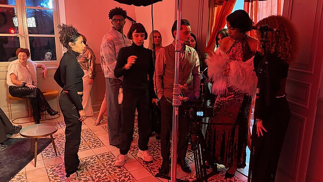 Cette image capture l'effervescence des coulisses d'un tournage de film dans une pièce élégante aux murs clairs et au sol recouvert de carreaux de ciment à motifs gris et blancs. L'ambiance est marquée par une lumière rouge intense qui baigne toute la scène, créant une atmosphère dramatique et sophistiquée.