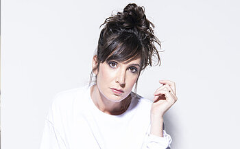 photo de l'humoriste Nora Hamzawi - L’image montre une femme photographiée sur un fond blanc uni. Elle a les cheveux bruns attachés en chignon haut avec quelques mèches tombant sur le front. Elle porte un haut blanc à manches longues. Son regard est dirigé vers la caméra, avec une expression neutre. Elle tient une main levée près de son visage, doigts légèrement repliés. L’éclairage est doux et uniforme.