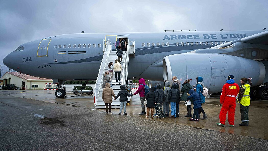 Cette image montre un groupe de personnes, dont plusieurs enfants, s'apprêtant à monter à bord d'un imposant avion militaire gris sur un tarmac mouillé. Un Aibus A330  MRTT, Il s'agit d'un avion de transport et de ravitaillement de l'armée de l'Air française, identifiable par l'inscription « ARMÉE DE L'AIR » sur le fuselage. Une file de personnes, vêtues de vêtements d'hiver (manteaux, bonnets), attend au pied de l'escalier. Certains enfants se tiennent par la main. Quelques personnes sont déjà en train de monter les marches de la passerelle. À droite, deux membres du personnel encadrent l'opération. L'un porte une combinaison rouge vif et l'autre un gilet de sécurité jaune fluorescent.