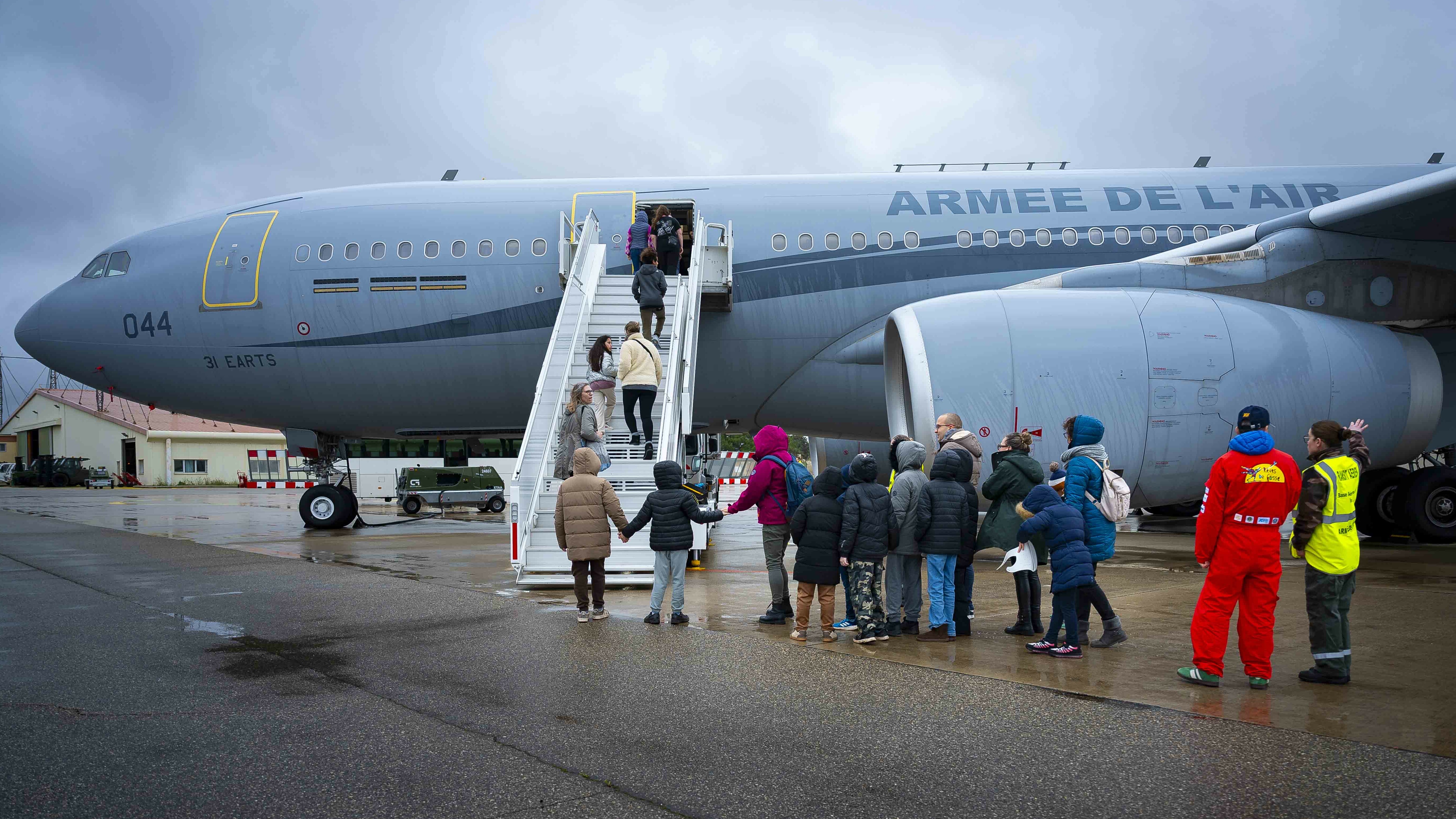 Cette image montre un groupe de personnes, dont plusieurs enfants, s'apprêtant à monter à bord d'un imposant avion militaire gris sur un tarmac mouillé. Un Aibus A330  MRTT, Il s'agit d'un avion de transport et de ravitaillement de l'armée de l'Air française, identifiable par l'inscription « ARMÉE DE L'AIR » sur le fuselage. Une file de personnes, vêtues de vêtements d'hiver (manteaux, bonnets), attend au pied de l'escalier. Certains enfants se tiennent par la main. Quelques personnes sont déjà en train de monter les marches de la passerelle. À droite, deux membres du personnel encadrent l'opération. L'un porte une combinaison rouge vif et l'autre un gilet de sécurité jaune fluorescent.