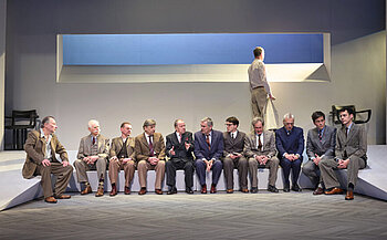 photo de la pièces de théâtre 12 hommes en colère  - L’image montre treize hommes dans une pièce aux murs clairs. Douze d’entre eux sont assis en ligne sur un banc blanc. Ils portent tous des costumes de couleurs variées, majoritairement sobres. Certains discutent entre eux, d’autres regardent droit devant. Le treizième homme, debout à l’arrière, regarde par une ouverture rectangulaire dans le mur, laissant apparaître un fond bleu. Deux chaises noires sont visibles en arrière-plan, à gauche et à droite. Le sol est gris avec un motif en chevrons.