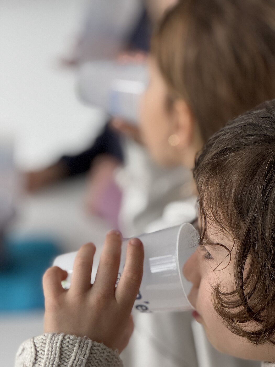 Cette photo montre deux enfants vus de profil, en train de boire dans des gobelets en plastique blanc.      Au premier plan : On voit le visage d'un enfant aux cheveux bruns bouclés, tenant fermement son verre avec sa main gauche. Il porte un pull en maille claire.      À l'arrière-plan : Un deuxième enfant, dont le visage est flou à cause de la faible profondeur de champ, effectue le même geste.      L'ambiance : L'éclairage est doux et le décor est minimaliste et lumineux, ce qui suggère un moment calme...