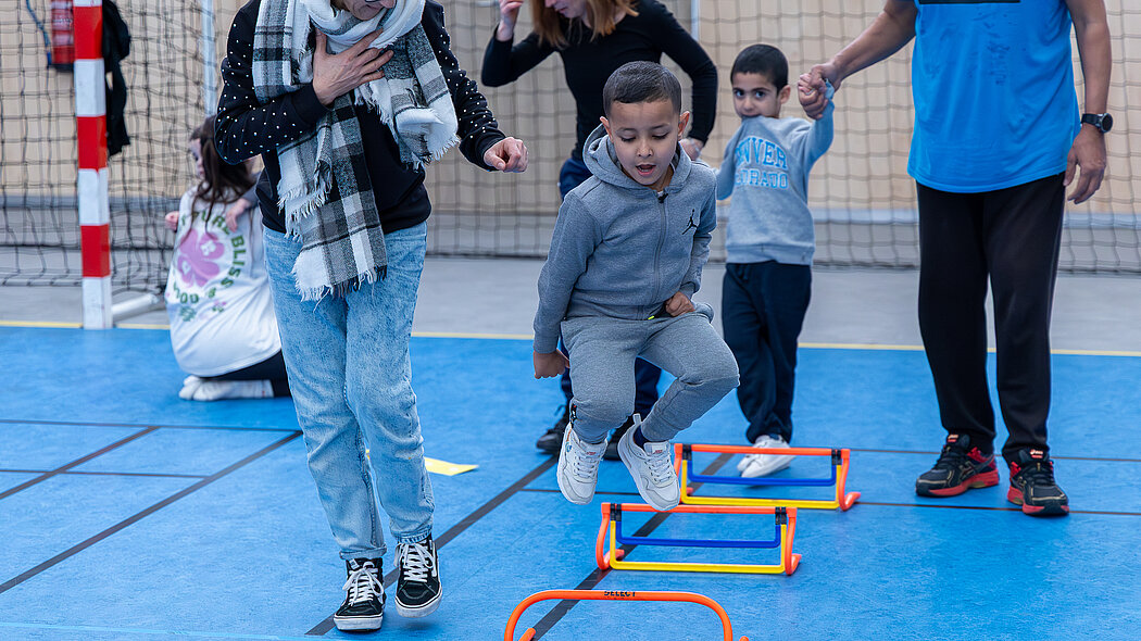 Cette image capture un moment dynamique et chaleureux lors d'un parcours de motricité pour enfants dans un gymnase au sol bleu.      L'action centrale : Un jeune garçon en survêtement gris est photographié en plein saut au-dessus de petites haies d'entraînement colorées.      L'accompagnement : Une femme à ses côtés (probablement une parente ou une éducatrice) l'accompagne et l'encourage avec le sourire.      À l'arrière-plan : Un autre enfant attend son tour en tenant la main d'un adulte en tenue de sport bleue, tandis que d'autres participants observent la scène dans une ambiance familiale et bienveillante.