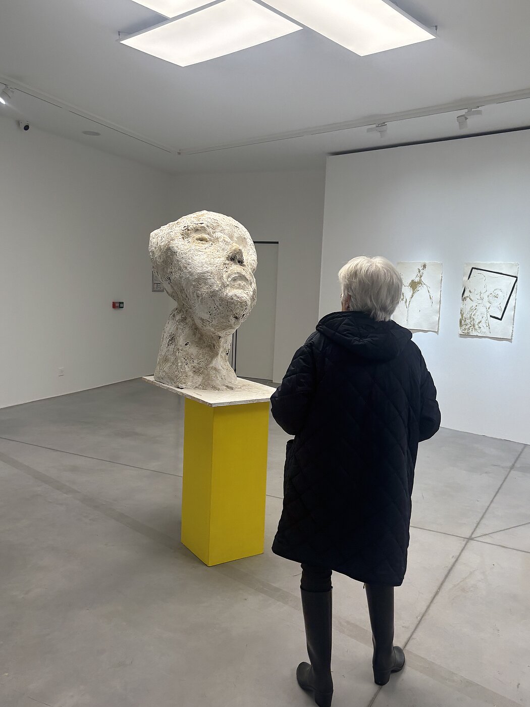 Cette image montre l'intérieur d'une galerie d'art moderne ou d'un musée, baignée d'une lumière blanche et diffuse provenant de panneaux rectangulaires au plafond.  ### La Pièce Centrale  Au cœur de la composition se trouve une **sculpture imposante** représentant une tête humaine.  * **Style :** Elle présente une texture rugueuse, presque organique, évoquant le plâtre, la pierre brute ou le papier mâché. * **Expression :** Le visage est orienté vers le haut, les traits (yeux, nez, bouche) sont suggérés de manière abstraite et tourmentée, ce qui lui donne un aspect émotionnel fort. * **Présentation :** La tête repose sur un socle rectangulaire d'un **jaune vif**, qui contraste radicalement avec la blancheur immaculée des murs et le gris neutre du sol en béton.  ### L'Observatrice  Une personne de dos contemple l'œuvre. Elle porte un long manteau matelassé noir et des bottes sombres. Ses cheveux sont courts et blancs. Sa position permet de se rendre compte de l'échelle de la sculpture, qui semble presque aussi grande que le buste d'un adulte.  ### L'Espace de la Galerie  * **Arrière-plan :** Sur le mur de droite, on aperçoit deux œuvres graphiques encadrées, de style minimaliste, avec des traits sombres sur fond blanc. * **Ambiance :** L'espace est très épuré et minimaliste, ce qui met l'accent sur les formes et les textures des objets exposés.