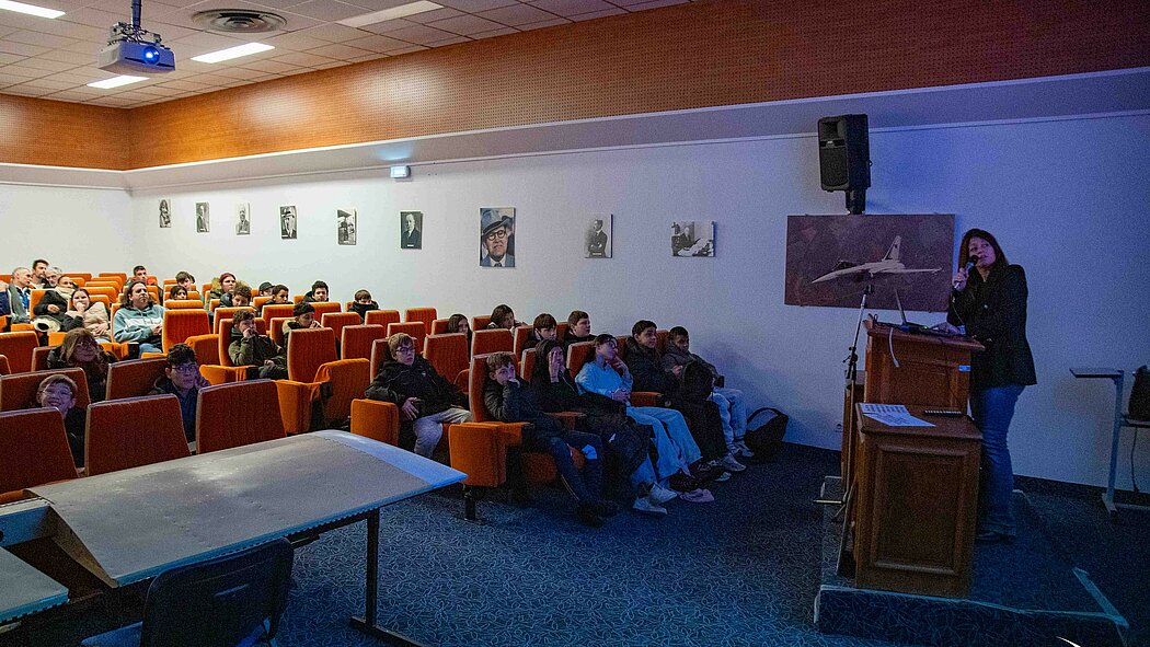 Cette photo offre une vue latérale de la salle, montrant davantage l'interaction entre l'intervenante et le public :      L'intervenante : Une femme se tient debout sur une estrade derrière un pupitre en bois, parlant dans un micro. Derrière elle, une affiche montre un avion de chasse en vol.      Le public : Un groupe de jeunes spectateurs est assis attentivement dans les rangées de sièges orange.      Le décor : Le mur latéral est orné d'une série de portraits photographiques en noir et blanc. Un vidéoprojecteur est fixé au plafond, diffusant de la lumière bleue vers l'écran
