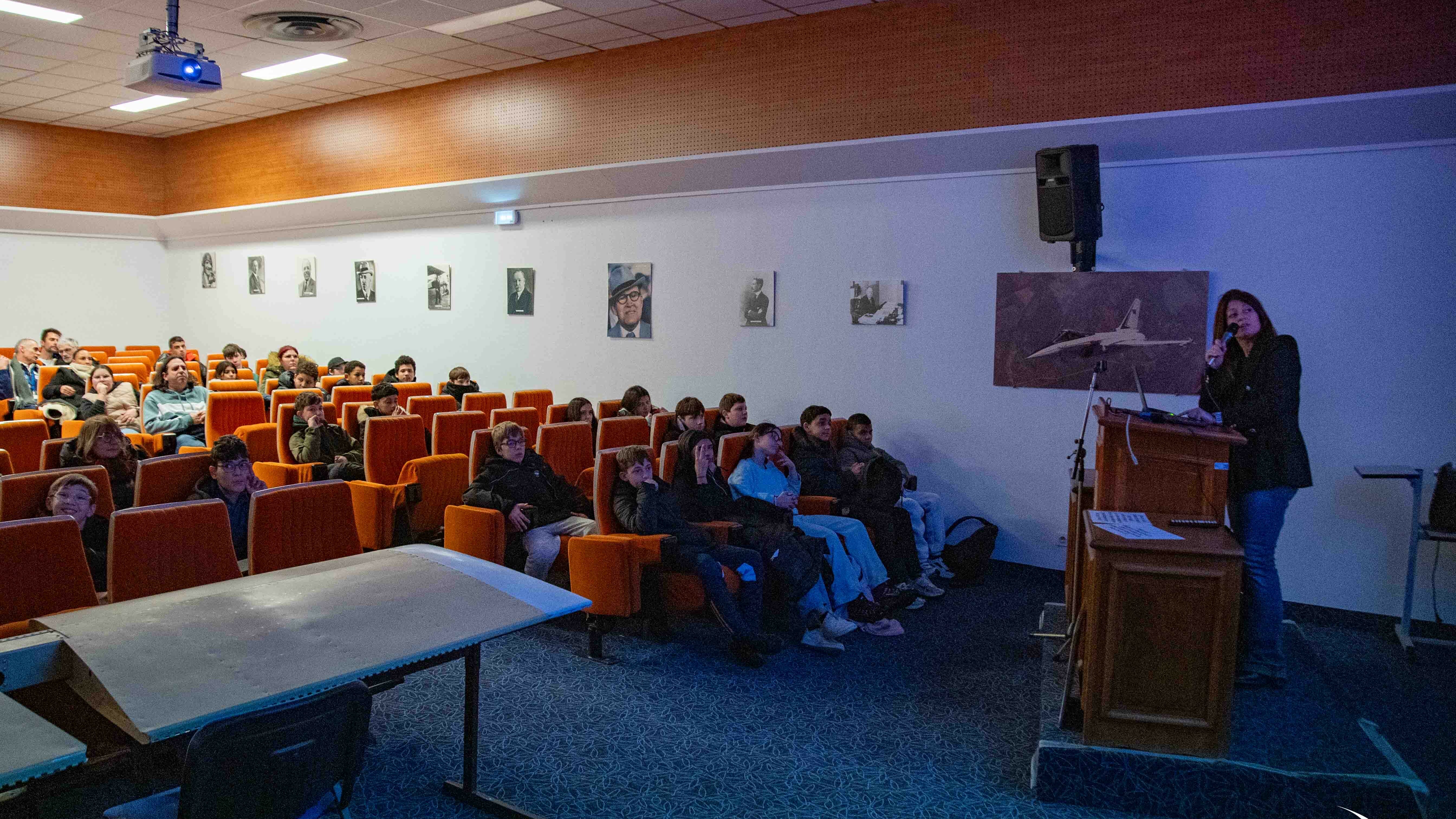 Cette photo offre une vue latérale de la salle, montrant davantage l'interaction entre l'intervenante et le public :      L'intervenante : Une femme se tient debout sur une estrade derrière un pupitre en bois, parlant dans un micro. Derrière elle, une affiche montre un avion de chasse en vol.      Le public : Un groupe de jeunes spectateurs est assis attentivement dans les rangées de sièges orange.      Le décor : Le mur latéral est orné d'une série de portraits photographiques en noir et blanc. Un vidéoprojecteur est fixé au plafond, diffusant de la lumière bleue vers l'écran