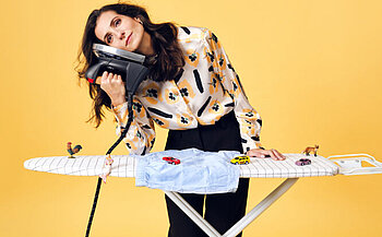 photo de l'humoriste Marine Leonardi  - L’image montre une femme debout devant un fond jaune uni. Elle porte un chemisier clair à motifs floraux jaunes et noirs, ainsi qu’un pantalon noir. Elle tient un fer à repasser contre sa joue, comme un téléphone. Devant elle se trouve une table à repasser recouverte d’une housse blanche à carreaux noirs. Sur la table sont posés de petits jouets, dont des voitures miniatures et des figurines d’animaux. Un vêtement bleu clair est également disposé sur la table.