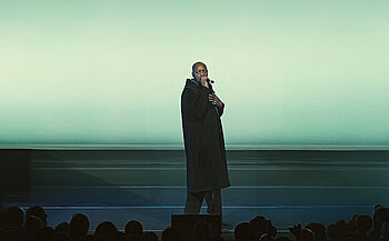 photo de l'artiste Oxmo Puccino - L’image montre une personne debout sur une scène, tenant un microphone dans sa main droite et plaçant sa main gauche sur sa poitrine. Elle porte un long manteau noir. Le fond de la scène est uniformément éclairé d’un dégradé vert clair. Des silhouettes de spectateurs sont visibles au premier plan, en bas de l’image. L’éclairage est centré sur la personne, mettant en valeur sa présence sur scène.