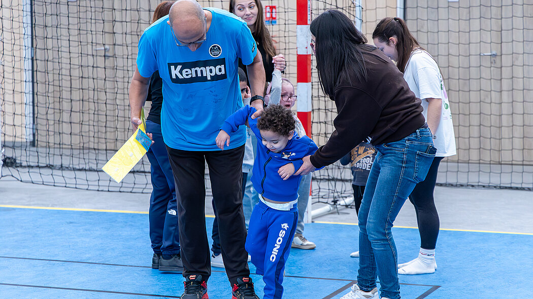 Cette image illustre un moment d'accompagnement bienveillant lors d'une activité d'éveil sportif dans un gymnase.      L'action centrale : Un jeune enfant, vêtu d'un survêtement bleu Sonic, réalise un exercice de motricité consistant à marcher dans des cerceaux rouges posés au sol.      L'encadrement : Deux adultes — un homme en t-shirt de sport bleu Kempa et une femme en jean et sweat marron — l'entourent et lui tiennent fermement les bras pour sécuriser ses pas et l'aider à garder l'équilibre.      L'ambiance : À l'arrière-plan, d'autres parents ou éducateurs observent la scène avec attention devant un filet de but, soulignant l'esprit d'entraide et de partage de cette séance.