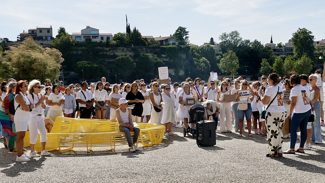 ette seconde photo montre le même rassemblement, mais sous un angle différent et plus large, mettant l'accent sur le moment du discours ou du recueillement.  Voici les éléments notables de cette image :      Le rassemblement : La foule, toujours vêtue majoritairement de blanc, s'est rassemblée en arc de cercle pour écouter une personne qui s'exprime au micro (à droite de l'image, de dos). L'atmosphère semble solennelle et attentive. Plusieurs personnes tiennent des fleurs blanches à la main en signe d'hommage.