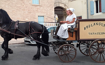 L’image montre une calèche tirée par un cheval noir. Deux femmes sont assises à l’avant de la calèche, habillées en tenues traditionnelles blanches avec coiffe. La calèche est en bois avec des roues à rayons, et porte l'inscription "BOULANGERIE TARASCON". À l’arrière-plan, on aperçoit des bâtiments anciens en pierre avec des volets bleus, ainsi qu’une voiture stationnée. La scène se déroule en milieu urbain.