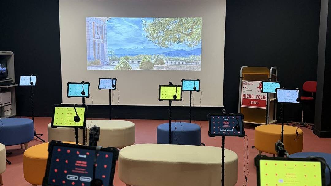 Salle MICRO FOLIE Salle intérieure avec un plafond noir et un éclairage LED en carrés.  Un grand écran de projection affiche une image de paysage (jardin, ciel bleu, arbre, bâtiment).  Plusieurs tablettes sont fixées sur des supports noirs devant l'écran.  Les tablettes affichent des écrans colorés (vert, bleu, rouge).  Plusieurs assises arrondies (poufs ou bancs) de différentes couleurs (jaune, beige, bleu) sont disposées dans la salle.