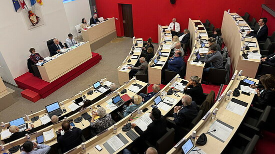 Photo de la salle de conseil municipal d'Istres - L’image montre une salle de réunion de type hémicycle, vue en plongée. Voici une description factuelle sans interprétation :     • La salle comporte plusieurs rangées de sièges disposées en demi-cercle, toutes orientées vers un bureau principal en hauteur, situé au fond de la pièce.     • Plusieurs personnes sont assises aux tables, munies d’ordinateurs portables, de tablettes, de documents papiers et de bouteilles d’eau.     • Sur l’estrade centrale, cinq personnes sont assises derrière un bureau surélevé, avec des micros.     • Au mur derrière cette estrade se trouvent quatre drapeaux (dont un drapeau européen, un drapeau français, et deux autres drapeaux régionaux).     • Un écran de projection est visible en haut à gauche, affichant un texte lisible partiellement.     • La pièce a des murs rouges et blancs, avec un sol de couleur beige et rouge.     • Il y a un espace vitré en hauteur, derrière lequel se trouvent deux personnes observant la scène.