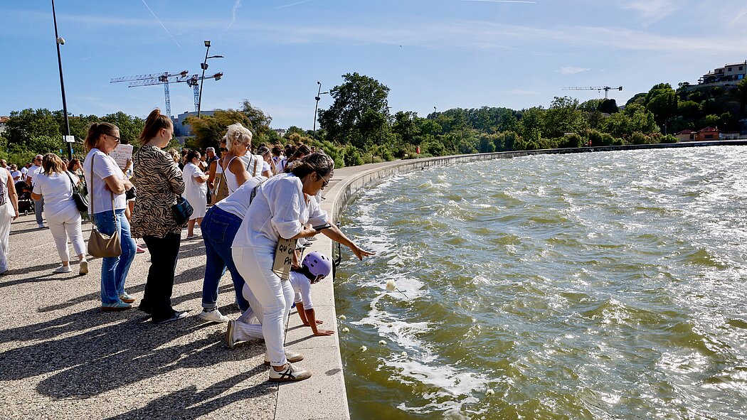 Cette photo capture le moment symbolique et émouvant du jet de fleurs dans l'eau.  Voici la description des éléments clés :      Le geste d'hommage : Au premier plan, on voit des participantes penchées au-dessus de la bordure en pierre. L'une d'elles, accompagnée d'un enfant portant un casque de protection violet, est en train de jeter une fleur blanche à l'eau. D'autres fleurs blanches sont déjà visibles à la surface, flottant sur l'écume.