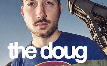 Photo du chanteur Jean + The Doug  - L’image montre un homme avec une barbe courte et des cheveux coupés courts, portant un t-shirt bleu. Il regarde droit vers l’objectif avec une expression neutre. En arrière-plan, une structure métallique inclinée est visible, suggérant un environnement urbain ou industriel. Le texte blanc en lettres minuscules superposées sur l’image indique « the doug ».