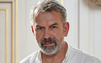 photo de l'acteur Philippe Torreton - L’image montre un homme aux cheveux gris et à la barbe grise, vêtu d’une chemise blanche légèrement ouverte au col. Il est placé devant un fond intérieur de couleur claire, avec des moulures murales décoratives en arrière-plan. L’éclairage est doux et met en valeur les traits de son visage.