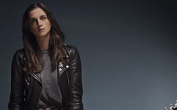 photo de la chanteuse Laura COX - L’image montre une femme aux cheveux longs et bruns, debout devant un fond uni bleu-gris. Elle porte une veste en cuir noir avec des boutons métalliques et un t-shirt gris. Elle regarde directement vers l’objectif avec une expression neutre. La partie inférieure droite de l’image laisse entrevoir une guitare électrique, dont seules les mécaniques sont visibles.