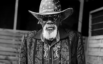 Photo du chanteur Robert Finley - L’image est en noir et blanc et montre une personne âgée, debout, regardant vers l’objectif. Elle porte un chapeau de style western orné d’étoiles, des lunettes de soleil, une veste à motifs floraux et une chemise brodée. La personne a une barbe blanche fournie. En arrière-plan, on distingue une structure en bois et une grille métallique sur un mur, suggérant un environnement extérieur ou semi-ouvert.