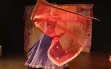 L'image montre une danseuse en train de performer sur scène. Elle porte un costume traditionnel composé d'une jupe bleue et d'un haut assorti, et elle tient un voile rouge translucide qu'elle fait virevolter autour d'elle. La scène est sombre, mettant en valeur la danseuse et son mouvement fluide. En arrière-plan, des motifs colorés, probablement des tissus décoratifs, sont visibles. Le voile crée un effet visuel dynamique et donne un sens de légèreté et de grâce à la danse.