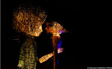 L’image montre une sculpture ou une installation artistique éclairée dans l'obscurité. Elle représente une figure humaine de profil, tournée vers la droite. Cette figure semble être fabriquée à partir de matériaux naturels tressés (comme de l'osier ou du rotin). La tête a une texture épaisse, évoquant des brindilles ou des fibres denses. Le bras droit de la figure est tendu vers un objet perché sur un bâton, qui ressemble également à une structure tressée, avec des fibres s'étendant vers l’extérieur.  La lumière provient de l’intérieur ou derrière les structures, créant un effet de transparence et mettant en valeur la texture des matériaux. Les couleurs dominantes de l’éclairage sont le jaune-orangé chaud et une touche de bleu sur le bâton central.  Crédit photo : © Sébastien CARLIER / PNRLG.