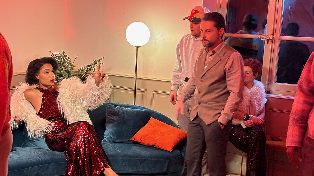 Cette image montre un moment de préparation sur un plateau de tournage, dans une atmosphère à la fois glamour et studieuse. La scène est baignée d'une lumière chaude et rosée qui provient de projecteurs hors champ, créant une ambiance feutrée de club ou de salon de luxe. L'ensemble de l'image suggère une grande attention portée aux détails esthétiques, typique d'une production cinématographique.