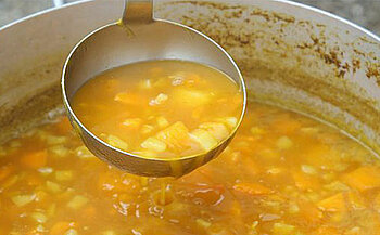 L’image montre un grand récipient métallique (marmite ou faitout) contenant un liquide de couleur orangée.  Des morceaux de légumes ou d’aliments de couleur jaune et orange flottent dans le liquide.  Une louche en métal est suspendue au-dessus de la marmite, remplie du même contenu.  Le bord intérieur de la marmite est taché ou coloré par des résidus du liquide.  En arrière-plan, on voit des jambes de personnes, l’une portant un pantalon blanc.  Le sol est en gravier ou cailloux.  Une poignée en métal est visible à l’avant de la marmite.