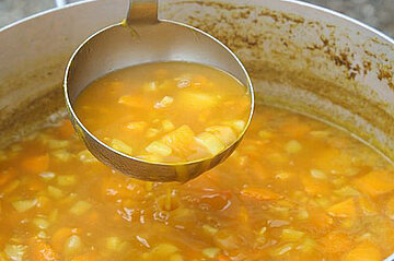 L’image montre un grand récipient métallique (marmite ou faitout) contenant un liquide de couleur orangée.  Des morceaux de légumes ou d’aliments de couleur jaune et orange flottent dans le liquide.  Une louche en métal est suspendue au-dessus de la marmite, remplie du même contenu.  Le bord intérieur de la marmite est taché ou coloré par des résidus du liquide.  En arrière-plan, on voit des jambes de personnes, l’une portant un pantalon blanc.  Le sol est en gravier ou cailloux.  Une poignée en métal est visible à l’avant de la marmite.