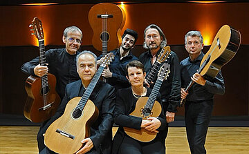 Sur cette image, six personnes posent en groupe sur une scène en bois, chacune tenant une guitare classique. Elles sont toutes habillées en noir, dans une tenue formelle. Trois personnes sont au premier plan, deux hommes et une femme, tenant leurs guitares de face. Trois autres sont debout à l’arrière, tenant également leurs instruments, dont l’un brandit sa guitare verticalement derrière sa tête.  Le fond est sombre, avec des lumières murales orange diffusant une lueur chaude. L’ambiance suggère un cadre de concert ou une répétition dans une salle dédiée à la musique. Les guitares sont toutes de style classique, en bois, avec des cordes en nylon.