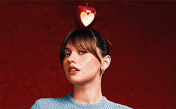 Crédit photo Elisa Baudoin - Marine de la star ac On voit une personne photographiée de face, cadrée du buste jusqu’à la tête. Elle porte un haut à manches longues de couleur bleu clair, avec une texture visible.  Ses cheveux sont châtain foncé, attachés en arrière, avec une frange qui tombe sur le front. Les oreilles sont visibles et portent de petites boucles d’oreilles.  La peau est éclairée de manière uniforme. Les lèvres sont légèrement entrouvertes. Le regard est dirigé vers l’avant.  Sur le sommet de la tête, il y a une pomme rouge. La pomme présente une découpe en forme de cœur laissant apparaître une partie plus claire à l’intérieur.  Le fond de l’image est uni, de couleur rouge foncé, avec un léger dégradé. La lumière est orientée vers le visage et le haut du corps, sans éléments visibles en arrière-plan.