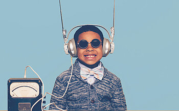 L’image montre un enfant souriant, portant un gros casque audio argenté avec deux antennes métalliques pointées vers le haut. Il porte également des lunettes rondes noires, un nœud papillon à rayures pastel, une chemise bleue et un cardigan texturé bleu. À sa gauche, un appareil analogique avec un cadran et des fils reliés au casque est posé sur une table. L’arrière-plan est uni et de couleur bleu clair.