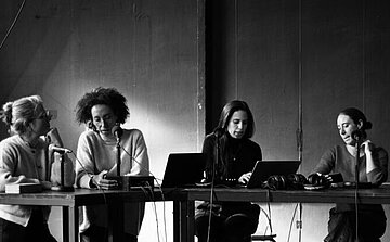 L’image en noir et blanc montre quatre personnes assises à une table. Chacune a un microphone devant elle. Deux ordinateurs portables sont posés sur la table, ainsi qu’un casque audio et divers objets. Les personnes sont tournées les unes vers les autres ou concentrées sur leurs ordinateurs ou leurs notes. Le mur en arrière-plan est sobre, sans décoration visible. Des câbles sont visibles sur et sous la table. L’ambiance est calme et studieuse.