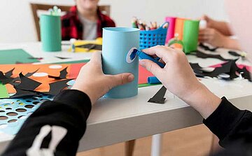 L'image montre un atelier de bricolage avec des enfants en arrière-plan. Un enfant est concentré sur l'assemblage d'un objet à partir d'un tube de papier bleu. L'enfant tient un œil en plastique et l'applique sur le tube à l'aide d'un bâton de colle. Sur la table, on peut voir plusieurs feuilles de papier de couleurs vives (orange, vert, bleu), des formes découpées en noir et en blanc, ainsi que des ciseaux et des outils de collage. Des crayons et des fournitures sont rangés dans un panier coloré à proximité, créant un environnement animé et créatif pour l'activité manuelle.