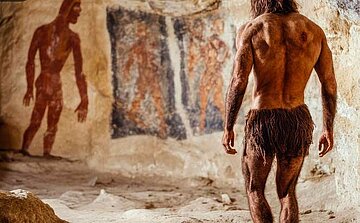 ChatGPT a dit :  L'image montre un homme debout, vu de dos, dans une grotte. Il porte une tenue primitive en peau, semblant être une sorte de pagne fait de fibres végétales. En arrière-plan, on distingue des peintures rupestres représentant des figures humaines, avec des formes stylisées et des couleurs rouges et noires. L'homme est musclé, et sa posture suggère qu'il observe les peintures. La scène semble se dérouler dans un environnement naturel, avec des murs de pierre apparents autour de lui.